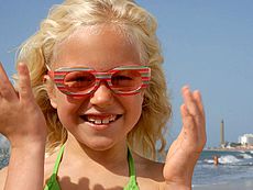 Flicka med färgade glasögon ler på stranden i Maspalomas med fyren som fond