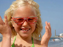 Lachendes Mädchen mit bunter Brille am Strand von Maspalomas, im Hintergrund der Leuchtturm