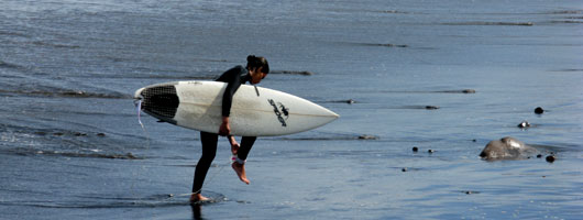 A girl walks along the water’s edge with a surf board under her arm