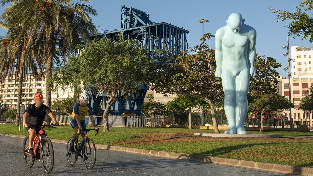 Two cyclists pass by Plaza de Canarias, where the Grúa Titán crane and the Greetingman sculpture are located in Las Palmas de Gran Canaria. Gran Canaria