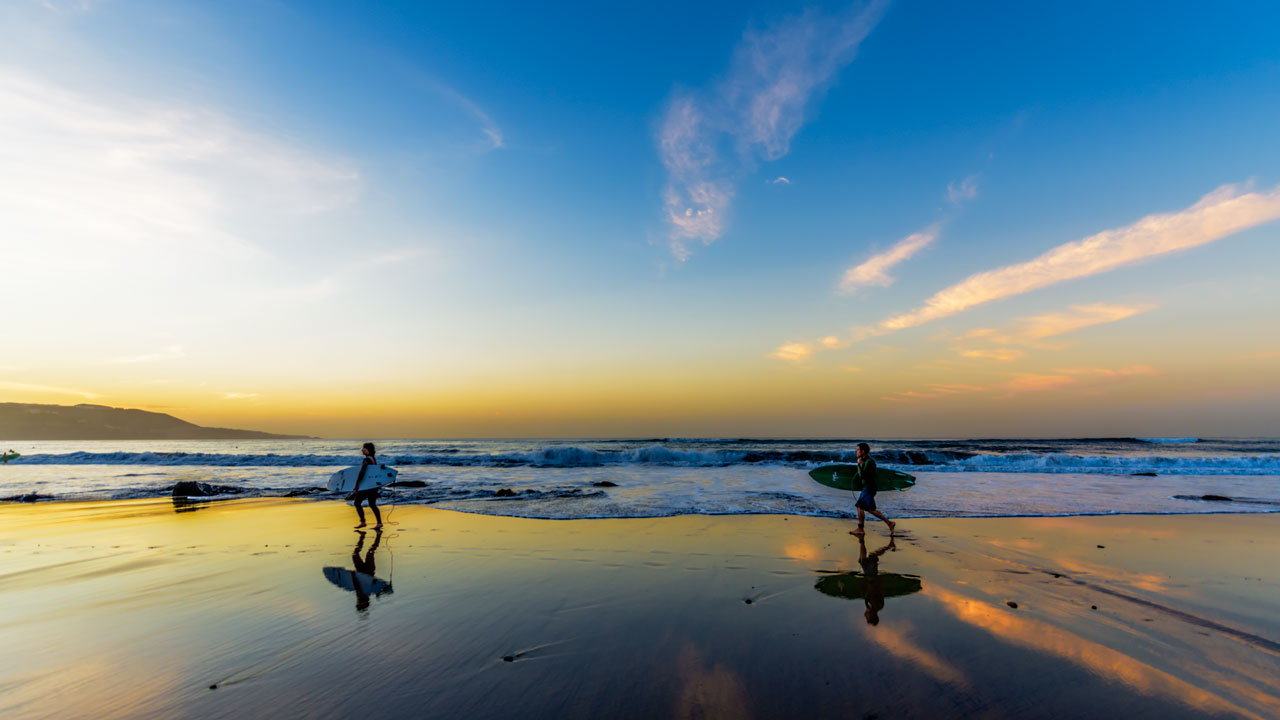 Surferos caminan por la orilla de la Playa de Las Canteras con sus tablas bajo el brazo, al atardecer